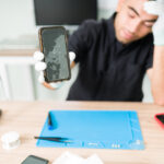 Stressed man repairing a broken old smartphone Look at this ugly smartphone. Upset male technician feeling worried while showing the cracked screen of a new smartphone