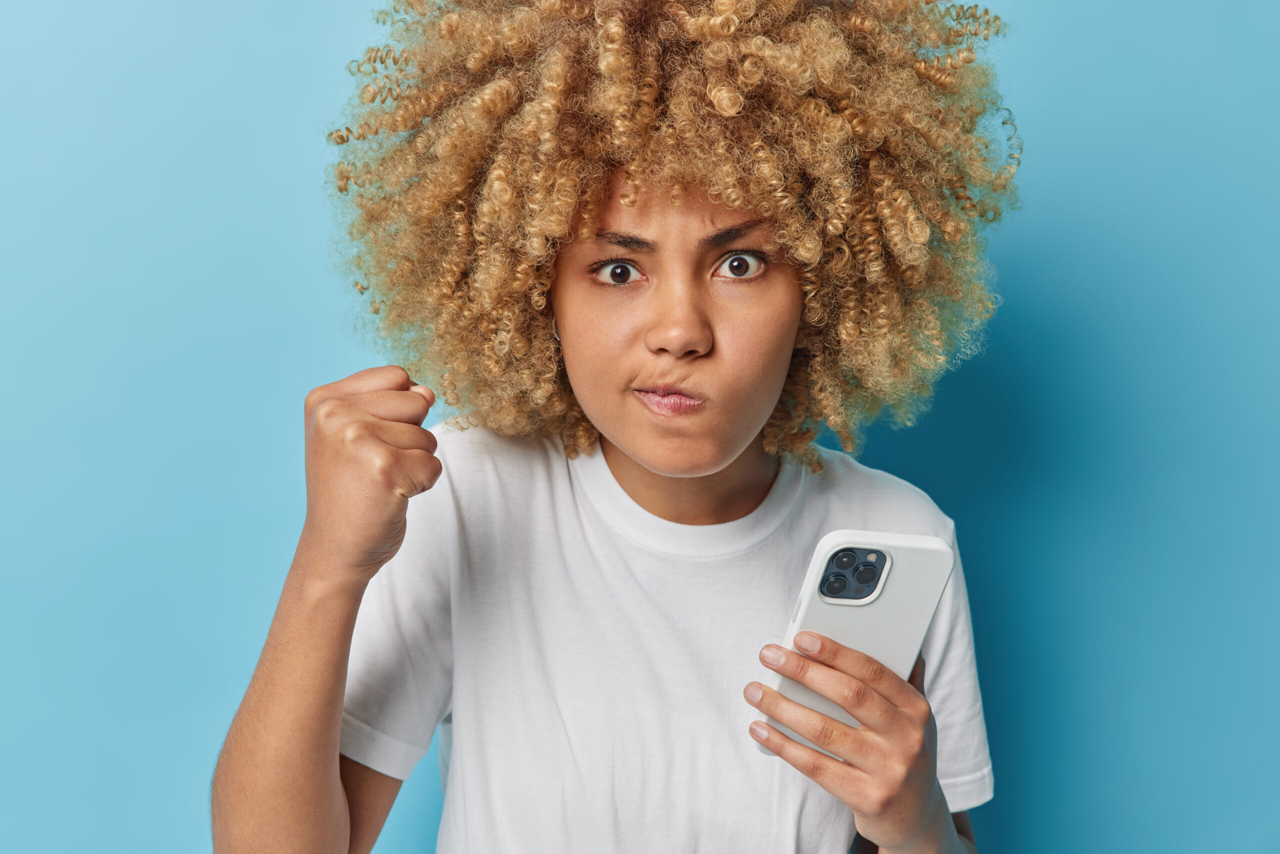 Photo of angry outraged curly haired woman purses lips clenches fist looks angrily uses modern mobile phone for sending text messages dressed in casual white t shirt isolated over blue background. Photo of angry outraged curly haired woman purses lips clenches fist looks angrily uses modern mobile phone for sending text messages dressed in casual white t shirt isolated over blue background.