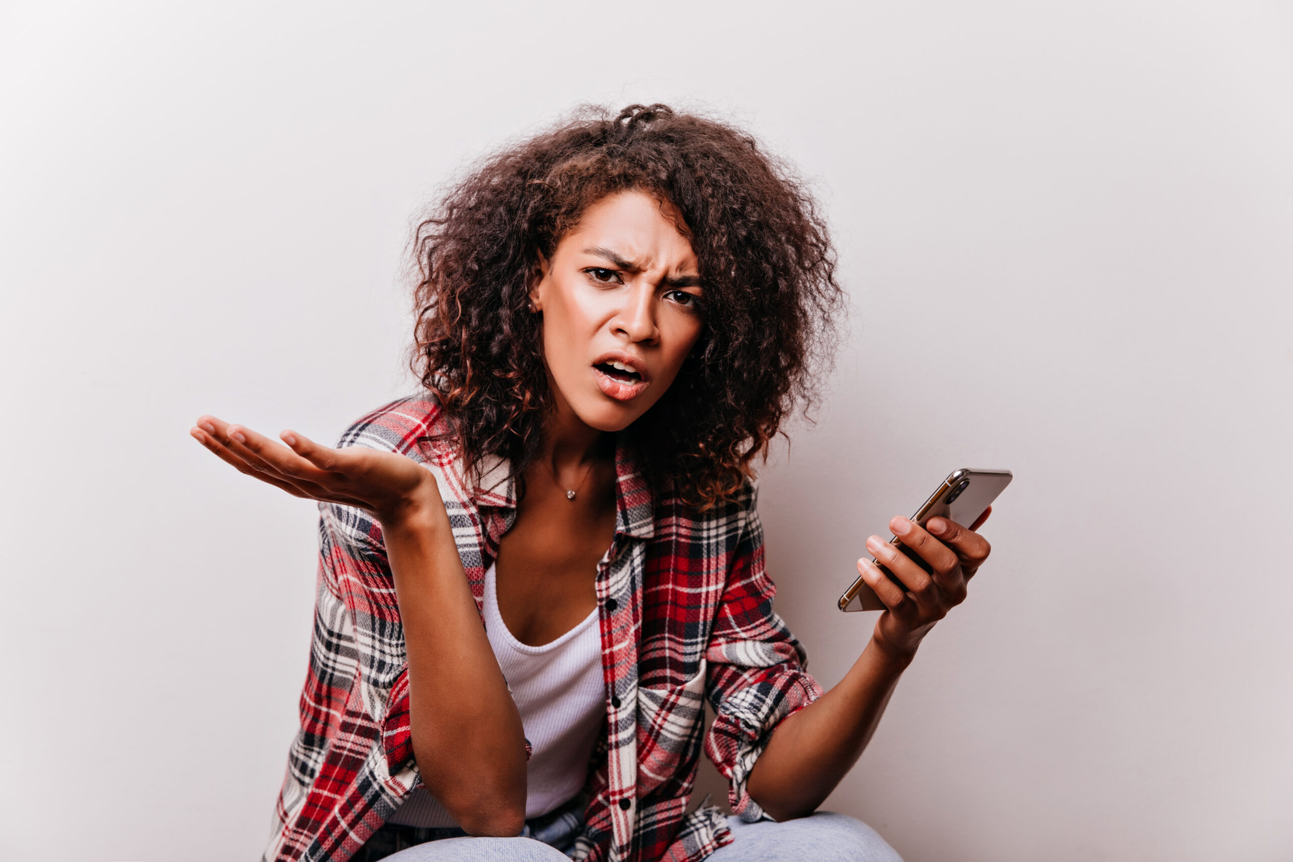 Unhappy black girl holding smartphone on white background. Surly Unhappy black girl holding smartphone on white background. Surly african lady in red shirt looking to camera.